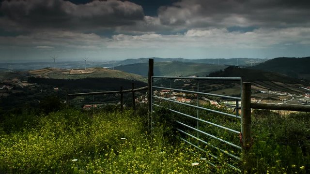 A static timelapse shot of the clouds rolling over a hilly landscape casting shadows above the wind turbines, while a fence sits on top of a hill in the foreground.