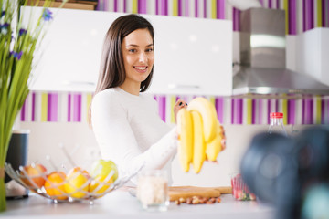 Beautiful and charming young woman is holding bananas in front of her kitchen counter filled with other fruits.