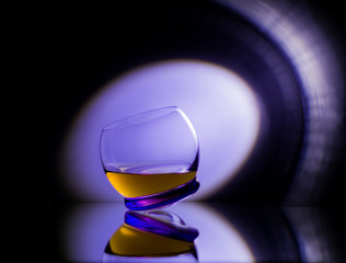 A bright round glass of a flip-flop with whiskey or juice on a black mirror surface with reflection in a ray of purple club light. Changed reality other condition at the party.