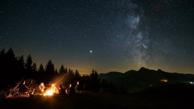 Friends sitting over campfire in starry night with milky way Time lapse