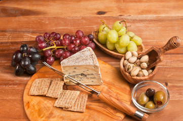 Top view, close, tapas, wedge of blue cheese, artisan cheese knife, crackers, rare, round wood plate, three varieties of grapes, wood bowl of pistachio nuts, and bowl of olives