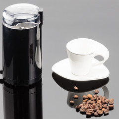 Coffee grinder with coffe beans and cup on the grey mirror background