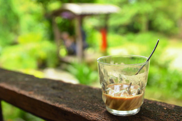 A glass of coffee with a spoon is placed on a wooden balcony in a garden atmosphere.