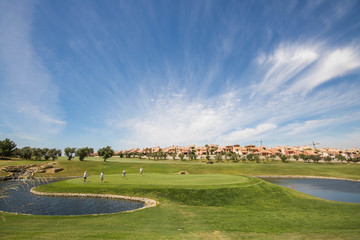 Four golfers playing at luxury golf course in Spain on a perfect summer day. Green surrounded by lakes with golf houses in the background.