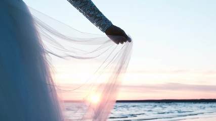 Woman hand in the lace sleeve holds the hem of the wedding dress. Transparent fabric fluttering in the wind at sunset against a background of a sea landscape in summer