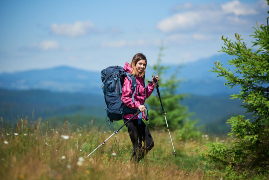 Young active female climber with backpack and trekking sticks, wearing sports wear, trekking in the mountains, smiling to the camera, enjoying sunny day. Concept of active lifestyle