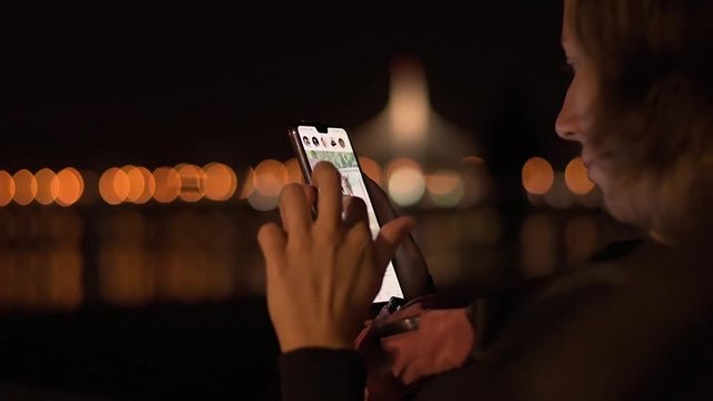 Young attractive lady sitting in the night park and wathing social media feed on her brand new mobile phone.