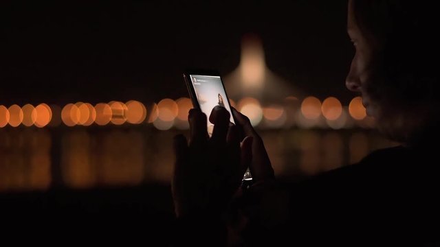 Young attractive lady sitting in the night park and wathing social media feed on her brand new mobile phone.