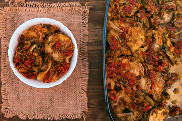 Glass dish with vegetable stew and one serving of dish laid out in white bowl, linen napkin, top view on wooden background.