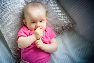 Fototapeta premium Sweet adorable baby girl puts fingers to mouth looking towards camera. 6-7 months old infant lying on back