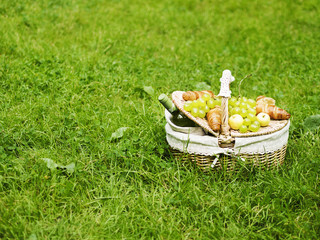 Wicker basket for picnic with green grape, apples, bottle of wine and croissants standing on green grass