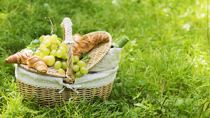 Wicker basket for picnic with green grape, apples, bottle of wine and croissants standing on green...