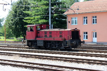 Used old dark red electric locomotive waiting for departure on railway tracks in front of new railway station building with bunch of wires above and trees in background