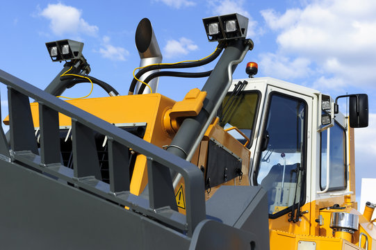 Bulldozer, huge yellow powerful construction machine with big grey scoop, heavy industry, white clouds and blue sky on background 