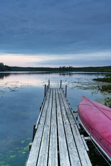 Red canoe tied to rustic wooden dock leading out to calm lake under overcast sunset sky