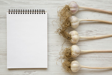Garlic bulbs with blank notepad on white wooden background, top view. Space for text.
