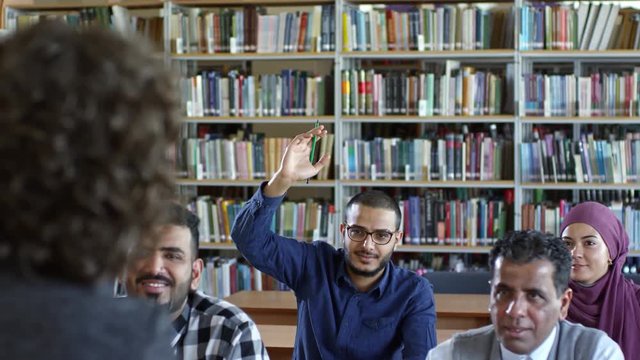 Tilt down of cheerful middle eastern man raising hand up and asking female teacher while studying language with group of migrant students in educational center