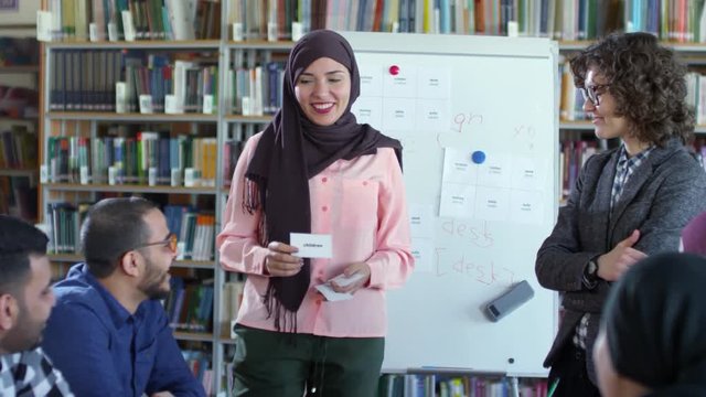 Beautiful muslim woman in hijab standing by flipchart in classroom, smiling and showing word cards to group of migrant students at English lesson with female teacher