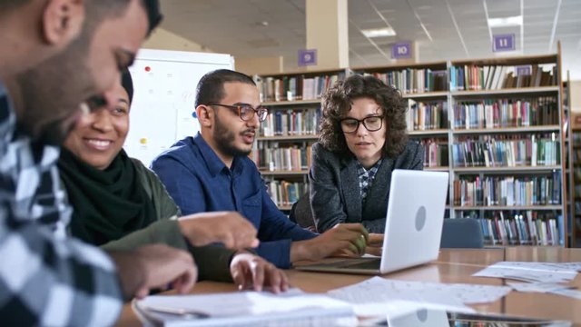 Black woman in hijab smiling and talking with middle eastern man at school desk while female teacher in eyeglasses explaining something on laptop screen to young arab man