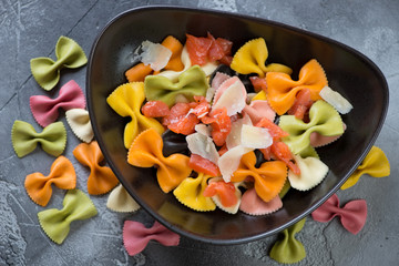 Black plate with multicolored farfalle, salmon and parmesan over grey concrete background, top view