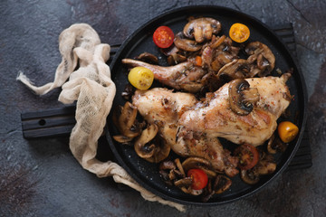 Metal tray with roasted rabbit meat, champignons and fresh cherry tomatoes. Flatlay over grey stone background