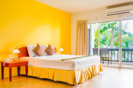 Interior decoration of romantic plain yellow bedroom with balcony. It consists of bed, white pillows, gold cushions, vintage lamps, telephone and side tables.