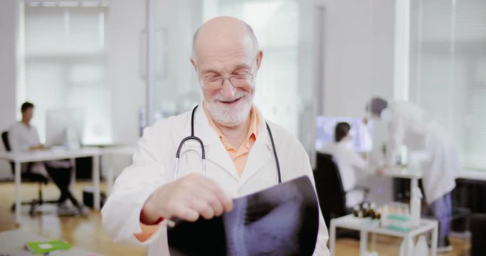 Elderly gray-haired man surgeon with glasses and stethoscope looking carefully x-ray of patient. Smiling doctor observes positive results of treatment