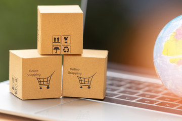 Stack of cardboard boxes with warning and shopping cart icon on laptop keyboard with globe nearby,...