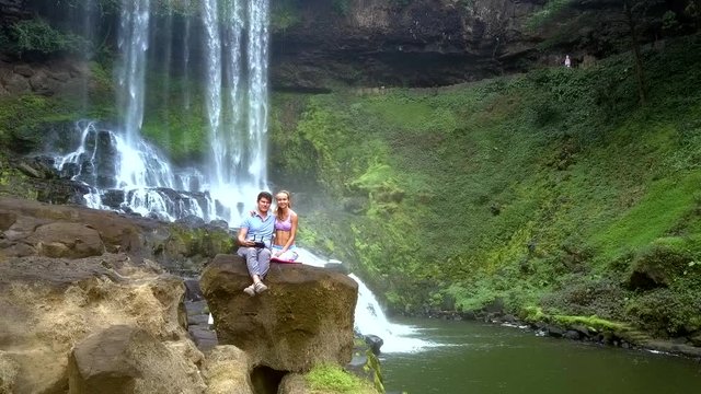 couple sits on rock guy controls flycam by waterfall