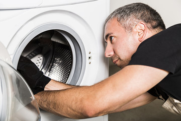 Plumber in uniform repairs drum of washing machine in laundry