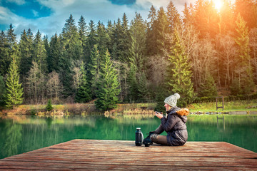 Happy young woman sitting on the wooden pier and drink coffee