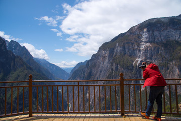 A tourist man standing on wooden long bridge & shooting photo  of beautiful view of big mountains in Balagezong national park in Shangri-La, China