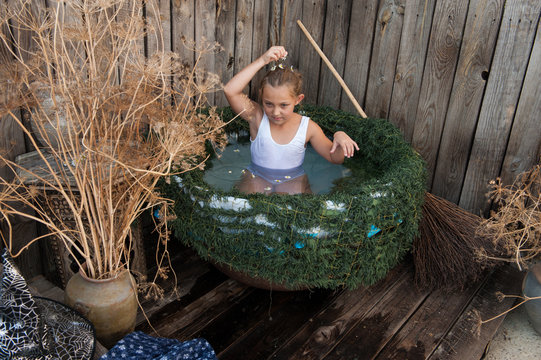 Young girl is preparing to take part in celebration of Halloween. Child is bathing in cauldron with chamomile flowers, trying on pointed hat. She is ready to fly on broomstick like a witch.