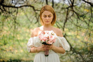 Happy blonde woman in a white dress with a bouquet on the background of the tree