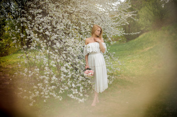Tender full height portrait od a blonde woman in white dress with a bouquet standing near the blooming tree on misty day