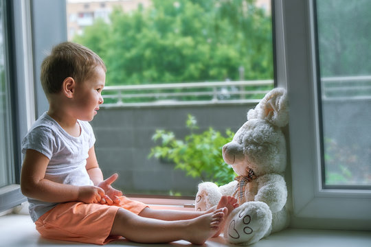 Boy Of Two Years Sitting By The Window And Hugs A Toy Bunny. Rainy Weather, Waiting For Dad To Come Home From Work.