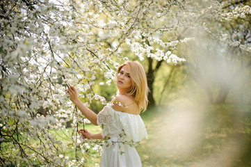 Portrait of lovely romantic blonde woman in white dress near the blooming tree