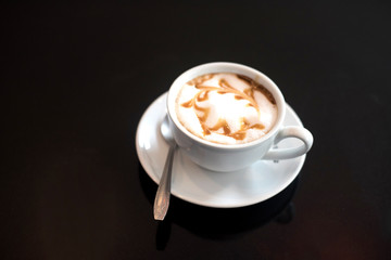 White cup of caramel coffee on black table for charge energy, refresh, breakfast or breaking time in afternoon. Cappuccino hot coffee on top with foam milk and caramel cream.