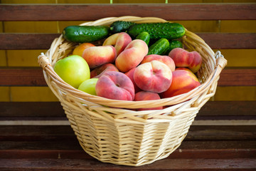 Woven from a vine basket stands on wooden boards filled with fruits from a fresh harvest. Peaches, apples, cucumbers.