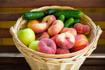 Woven from a vine basket stands on wooden boards filled with fruits from a fresh harvest. Peaches, apples, cucumbers.
