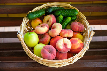 Woven from a vine basket stands on wooden boards filled with fruits from a fresh harvest. Peaches, apples, cucumbers.