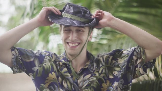 Young male traveller puts on his wide brim hat and stares into the camera. In tropical setting with Palm Trees