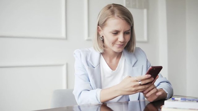 Beautiful and happy, successful business woman with mobile phone and notepad in a cafe, working as a freelancer. Blonde Female in blue formal suit sitting at the table and using mobile application