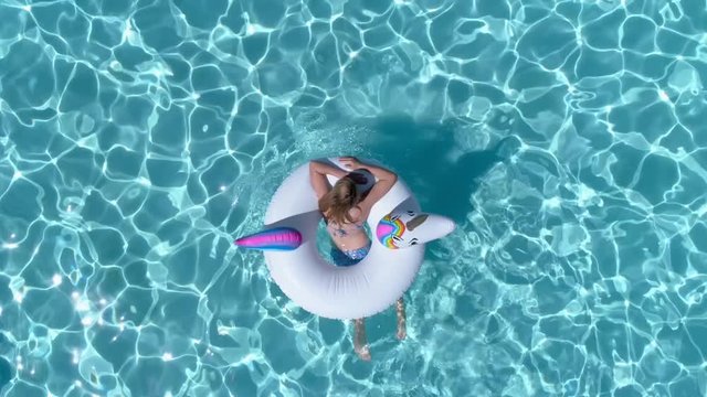 Top down view of a crystal blue swimming pool as a young girl enters into frame atop a unicorn shaped pool float.