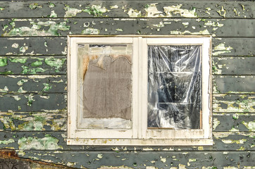 Window with broken glass, closed with plastic and plywood; in a wooden facade with peeling green paint