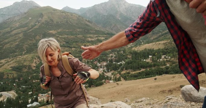 Mature caucasian senior on a hiking adventure taking wife's hand helping her climb up a mountain. tourism concept 4k