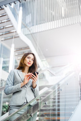 A young brunette girl in a gray jacket is standing in a bright corridor  looking into the distance and holding the phone. Business, technology and communication concept
