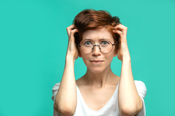 Frustrated or discontent red haired caucasian woman keeping arms on head being in dispare as if she did something irreparrable, looking at camera isolated on blue background