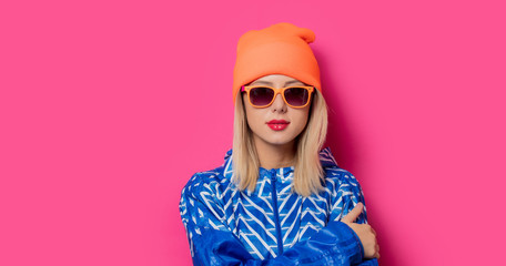 Young blonde girl in 90s sports jacket and hat with sunglasses on pink background.