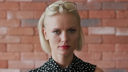Positive young female with stylish glasses on head smirking and looking at camera while standing on background of brick wall
